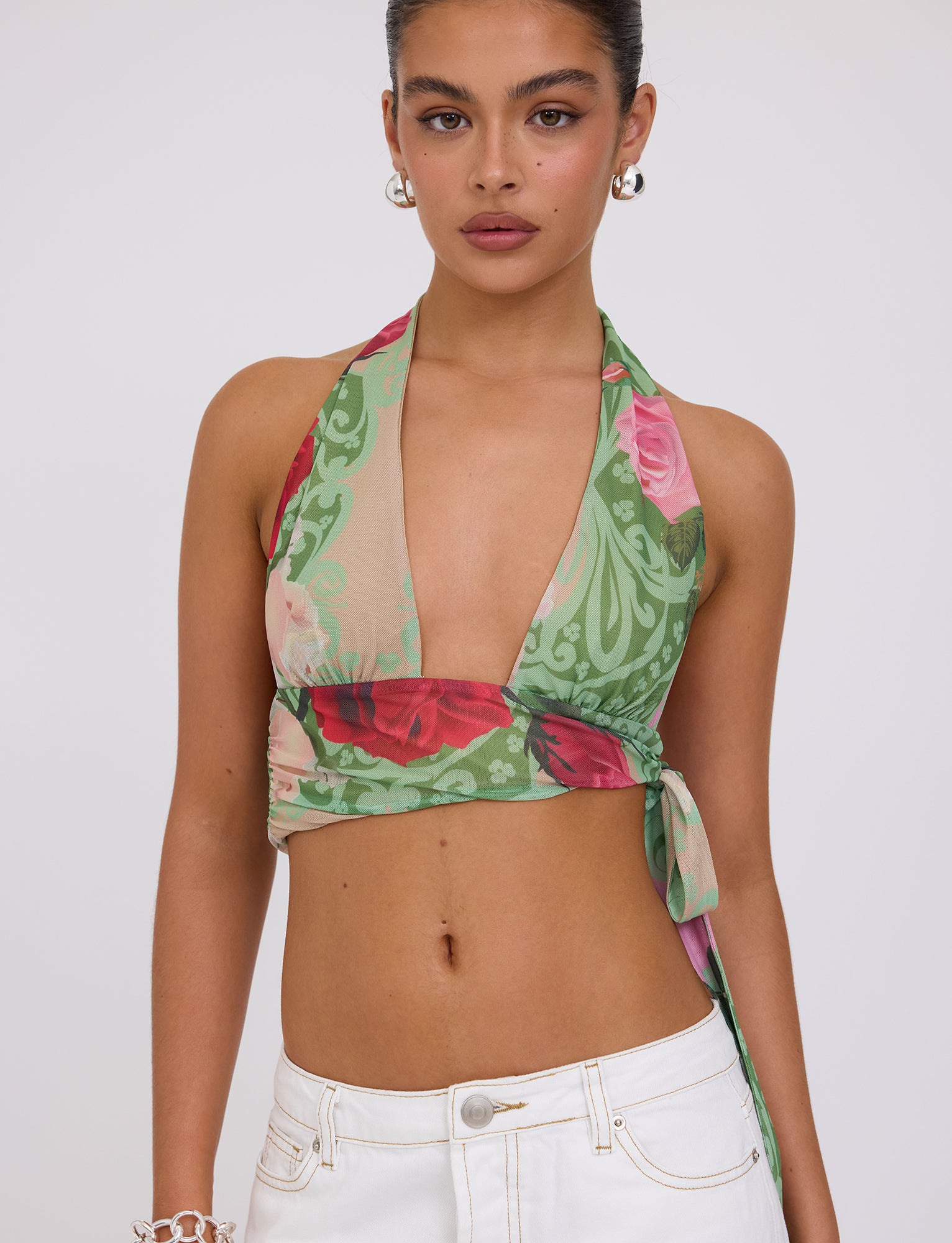 A woman with tied-back hair is wearing the Florian Top - Multi: Macro Floral: Summer Rose from Tigermist, which features a plunging neckline and green, red, and pink floral patterns. She also has large silver hoop earrings and white high-waisted pants. The background is plain white.
