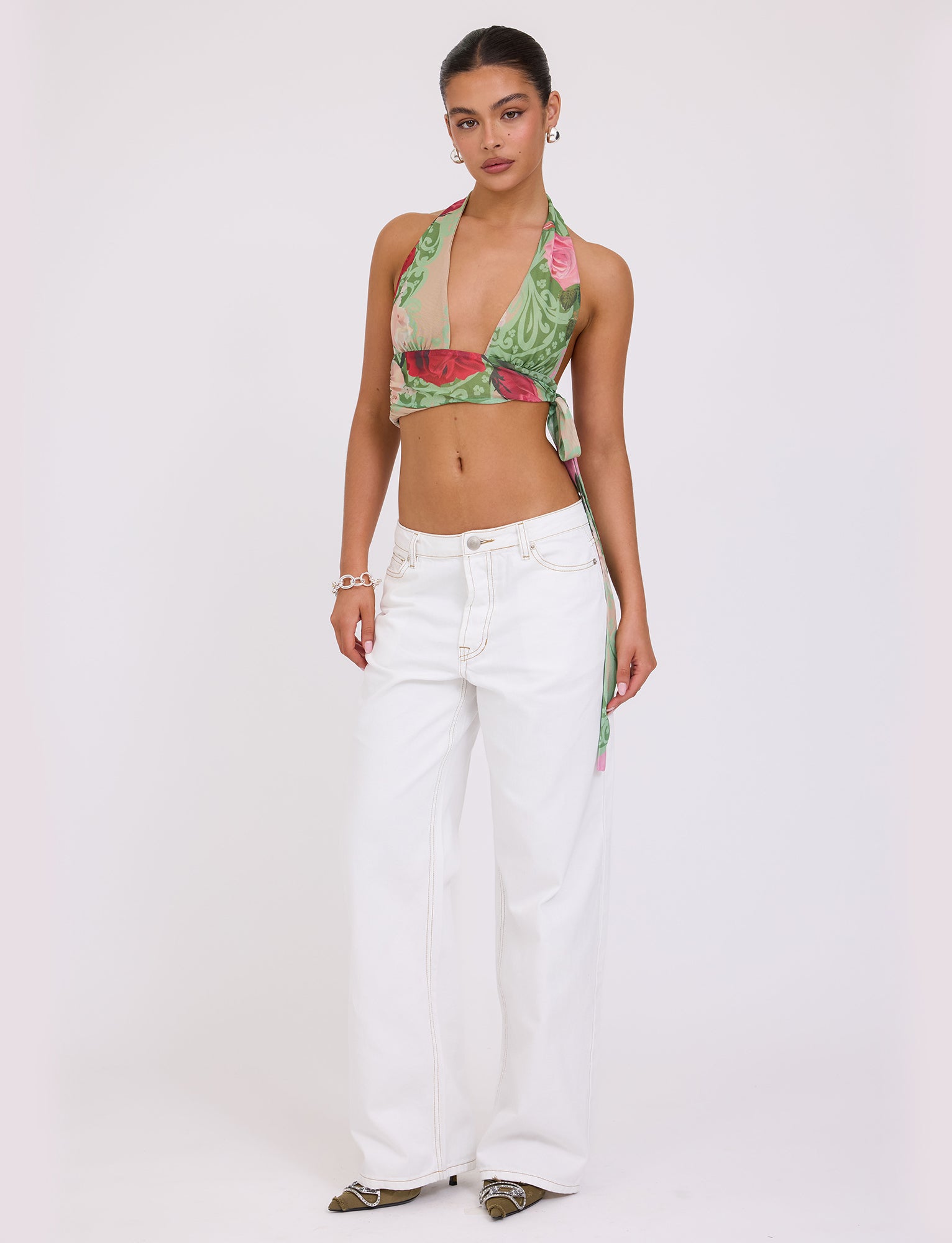 A person stands against a plain background wearing the FLORIAN TOP - MULTI : MACRO FLORAL : SUMMER ROSE from TIGERMIST, paired with white wide-leg pants. They accessorize with earrings, a bracelet, and pointed shoes. With a confident expression, they pose with one hand on their hip.