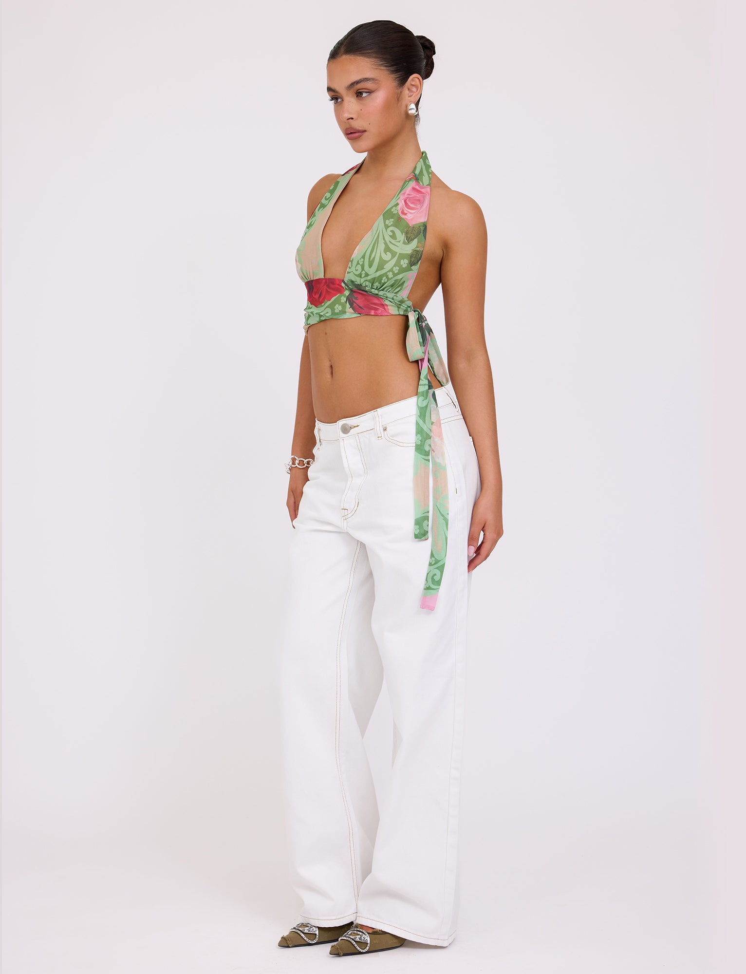 A person with dark hair styled in a bun is posing against a plain background, wearing the TIGERMIST FLORIAN TOP - MULTI : MACRO FLORAL : SUMMER ROSE halter top and wide-legged white jeans. They accessorize with bracelets and have a confident stance, gazing slightly to the side.