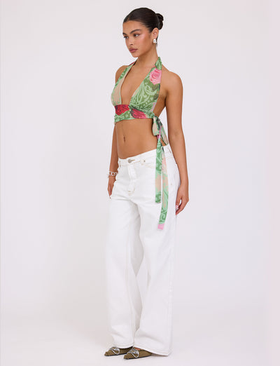 A person with dark hair styled in a bun is posing against a plain background, wearing the TIGERMIST FLORIAN TOP - MULTI : MACRO FLORAL : SUMMER ROSE halter top and wide-legged white jeans. They accessorize with bracelets and have a confident stance, gazing slightly to the side.