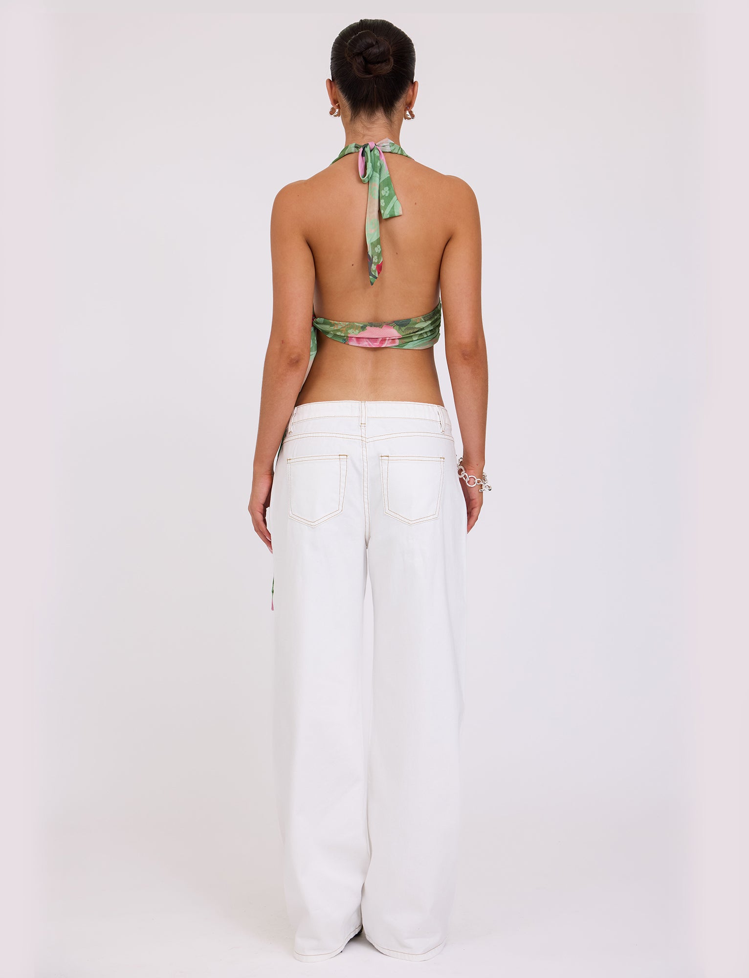 A person is standing with their back to the camera, wearing the Florian Top - Multi: Macro Floral: Summer Rose by TIGERMIST, which features a green floral halter design, paired with white wide-legged jeans. They have their hair styled in a bun and are accessorized with earrings and a bracelet.