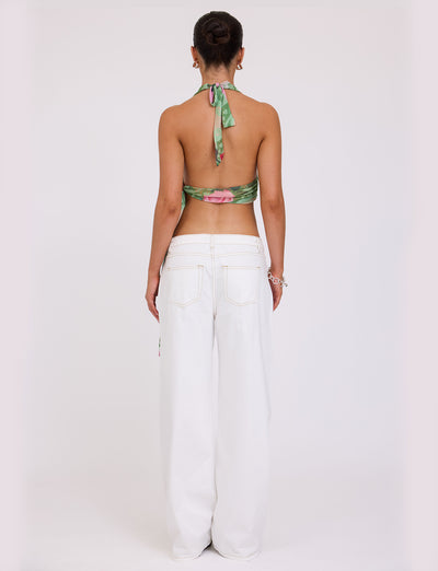 A person is standing with their back to the camera, wearing the Florian Top - Multi: Macro Floral: Summer Rose by TIGERMIST, which features a green floral halter design, paired with white wide-legged jeans. They have their hair styled in a bun and are accessorized with earrings and a bracelet.