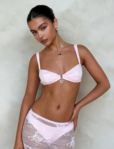 A woman is posing against a textured, neutral-colored wall, wearing a light pink Xadie Top by TIGERMIST featuring a heart-shaped charm in the center and a matching lace cover-up skirt. Her hair is tied back, and she accessorizes with a necklace and earrings.