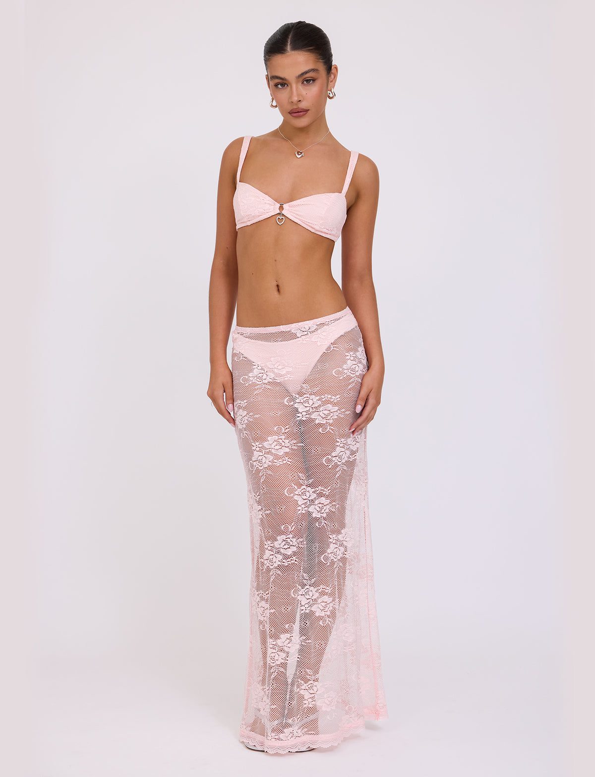 A woman stands against a plain background wearing the XADIE TOP in pale pink from TIGERMIST, paired with a matching sheer, floor-length lace skirt. She has long hair tied back and is accessorized with earrings and a necklace from Tiger Mist. She gazes directly into the camera with a neutral expression.