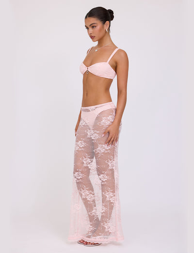 A woman stands against a plain background wearing the XADIE TOP from TIGERMIST in pastel pink. The outfit includes a bra, matching underwear, and a sheer lace skirt that extends to her feet. Her hair is styled neatly in a bun as she poses with her body turned slightly to the side.