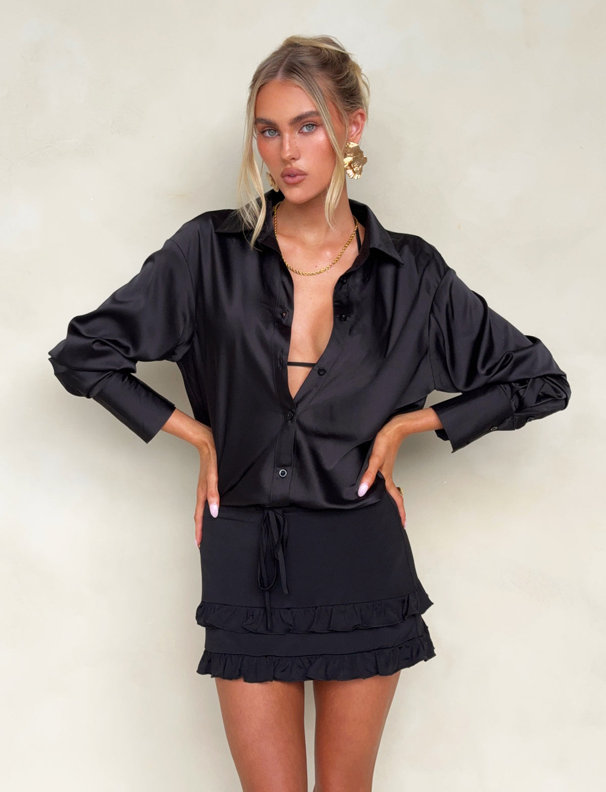 A woman with long blonde hair stands confidently, hands on her hips. She is wearing a silky black ALINA SHIRT from TIGERMIST, partially unbuttoned, paired with a short black skirt featuring ruffled hem detailing. A neutral background completes the image.