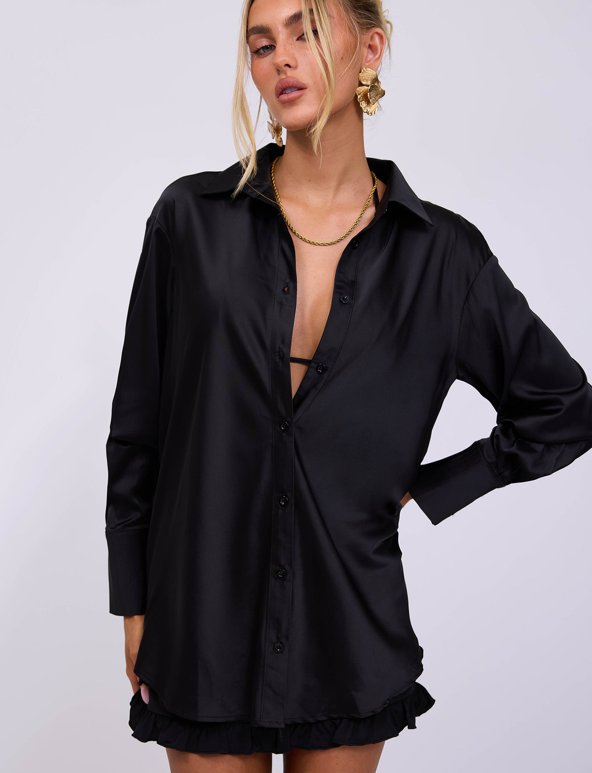A person with blonde hair is wearing the ALINA SHIRT - BLACK from TIGERMIST, a black, long-sleeved shirt with a deep neckline, along with gold statement earrings. The stylish shirt is paired with a black, ruffled mini skirt. They pose with one hand on their hip against a plain white background.