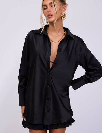 A person with blonde hair is wearing the ALINA SHIRT - BLACK from TIGERMIST, a black, long-sleeved shirt with a deep neckline, along with gold statement earrings. The stylish shirt is paired with a black, ruffled mini skirt. They pose with one hand on their hip against a plain white background.