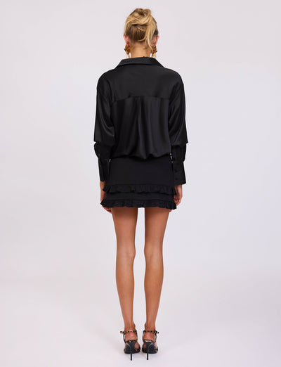 A person with blond hair stands facing away, wearing a chic TIGERMIST ALINA SHIRT in black and a short, ruffled black skirt. Their earrings glint under the light as they balance gracefully in black high-heeled shoes with decorative ankle straps. The background is plain white.