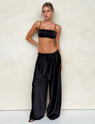 A woman stands against a light-colored wall, wearing the ALINA PANT - BLACK and a black spaghetti strap crop top from TIGERMIST. Her long blonde hair is styled in a sleek bun, and she accessorizes minimally with a bracelet on her right wrist. Her relaxed pose exudes confidence.