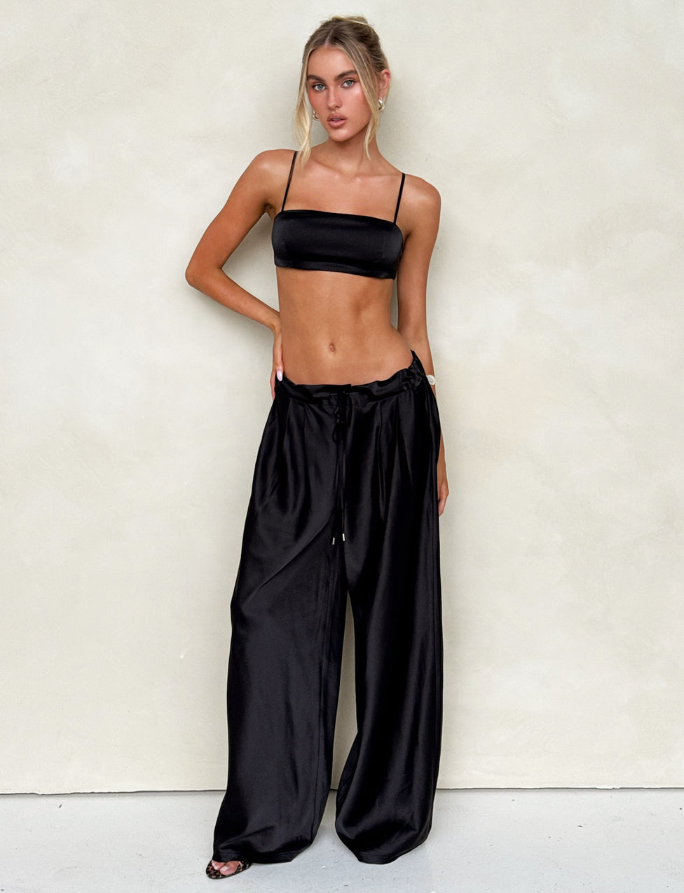 A woman stands against a light-colored wall, wearing the ALINA PANT - BLACK and a black spaghetti strap crop top from TIGERMIST. Her long blonde hair is styled in a sleek bun, and she accessorizes minimally with a bracelet on her right wrist. Her relaxed pose exudes confidence.