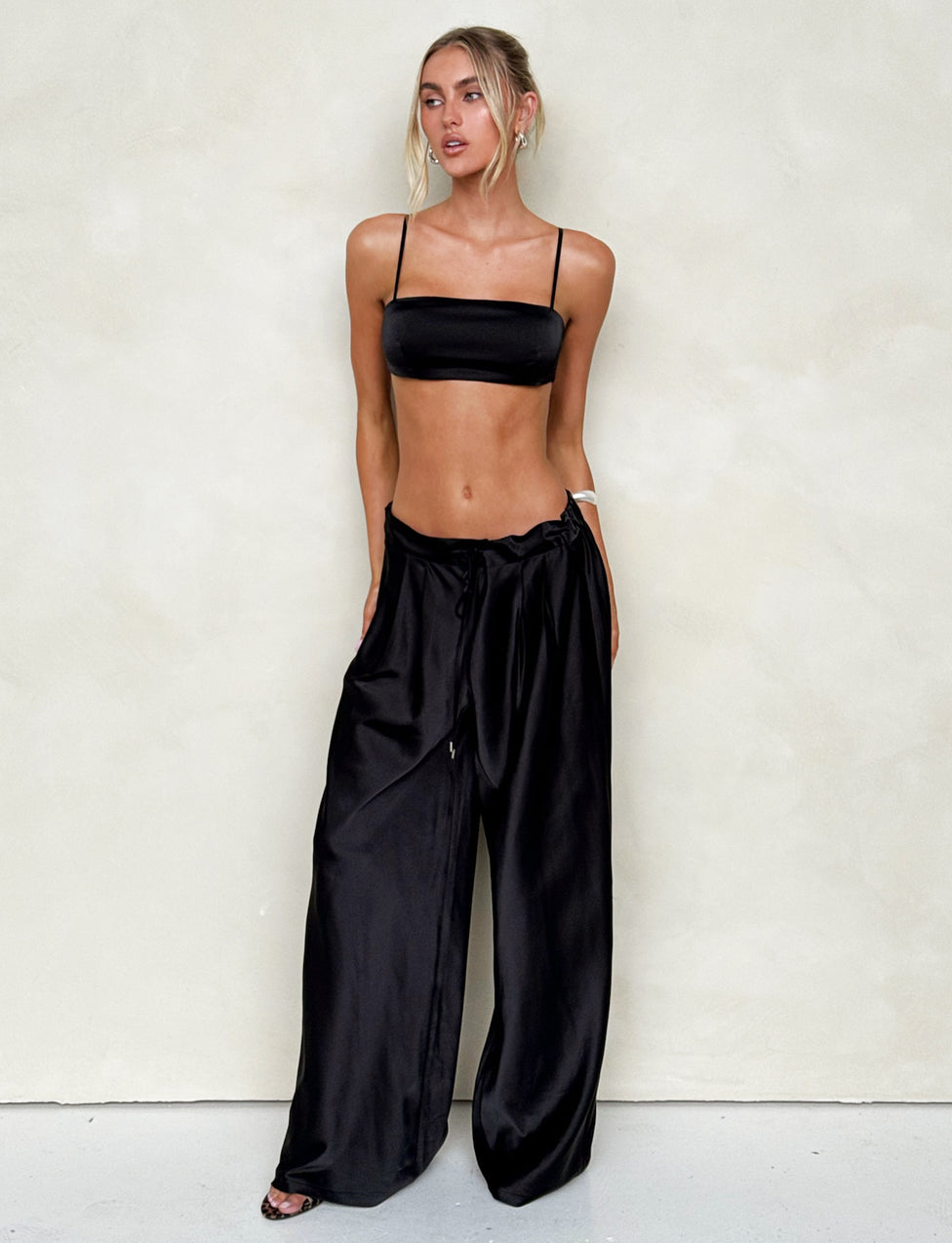 A woman stands against a plain light-colored background, wearing the sleek ALINA TOP - BLACK from TIGERMIST and high-waisted, wide-leg pants. She has blonde hair tied back with loose strands framing her face and is looking slightly to her right.