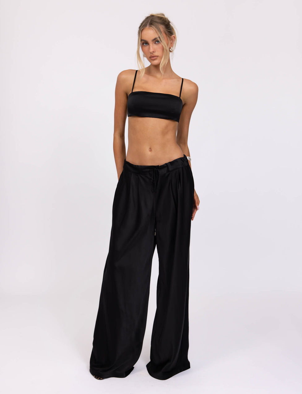 A person stands confidently against a plain background, sporting the TIGERMIST ALINA PANT in black along with a black bandeau top featuring thin straps. They have long blonde hair styled in a half-up, half-down fashion and maintain a relaxed posture.