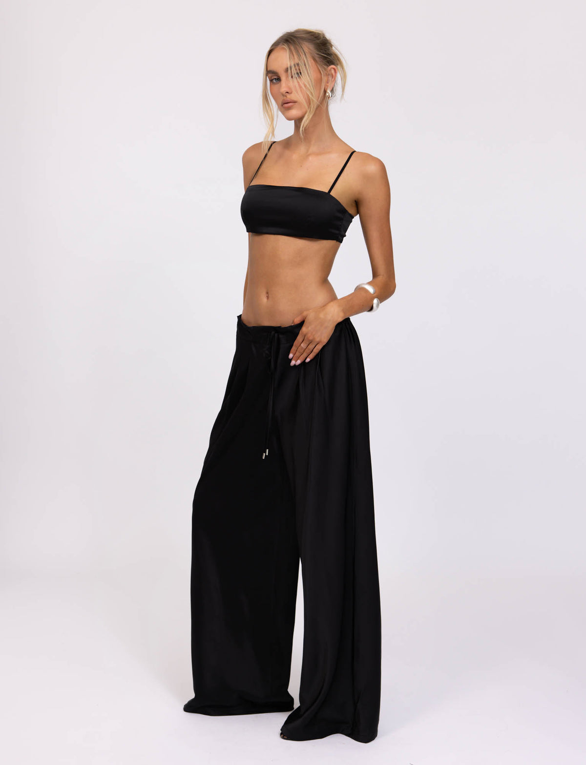 A woman stands against a plain white background wearing the chic TIGERMIST ALINA TOP in black, featuring thin straps, paired with loose-fitting black pants. Her hair is styled in a loose updo, with strands framing her face as she rests one hand on her hip and the other by her side.