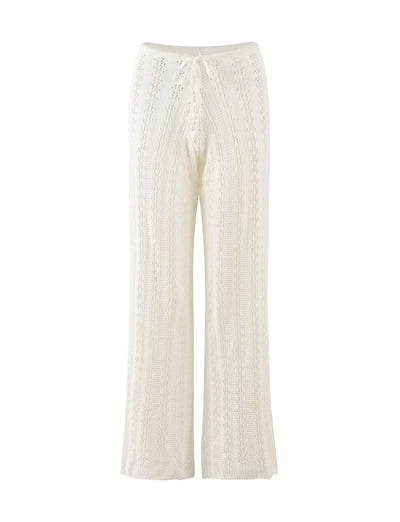 The SANDOR PANT - WHITE from TIGERMIST features a wide-leg design with a drawstring waist. Made from a white, knitted fabric with a subtle vertical cable-knit pattern, these pants offer both texture and a slightly sheer appearance. The hem falls just below the ankles.