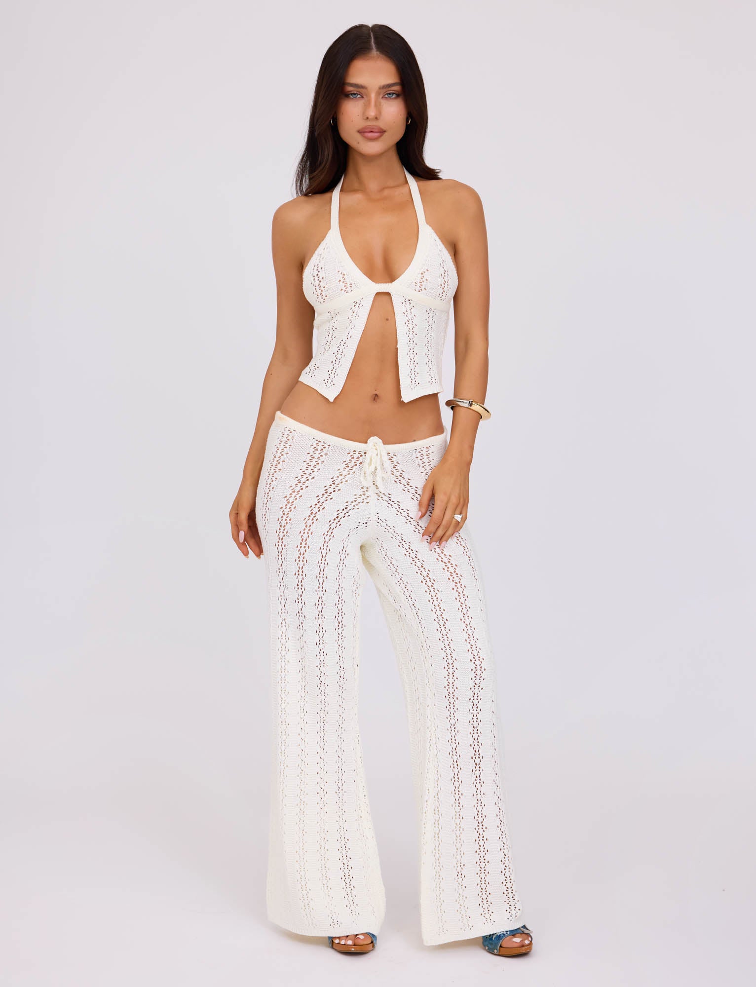 A woman with long dark hair is wearing a white crochet halter top and the SANDOR PANT - WHITE from TIGERMIST. She is standing against a plain light background with a neutral expression, her right hand resting on her hip while her left arm hangs by her side.