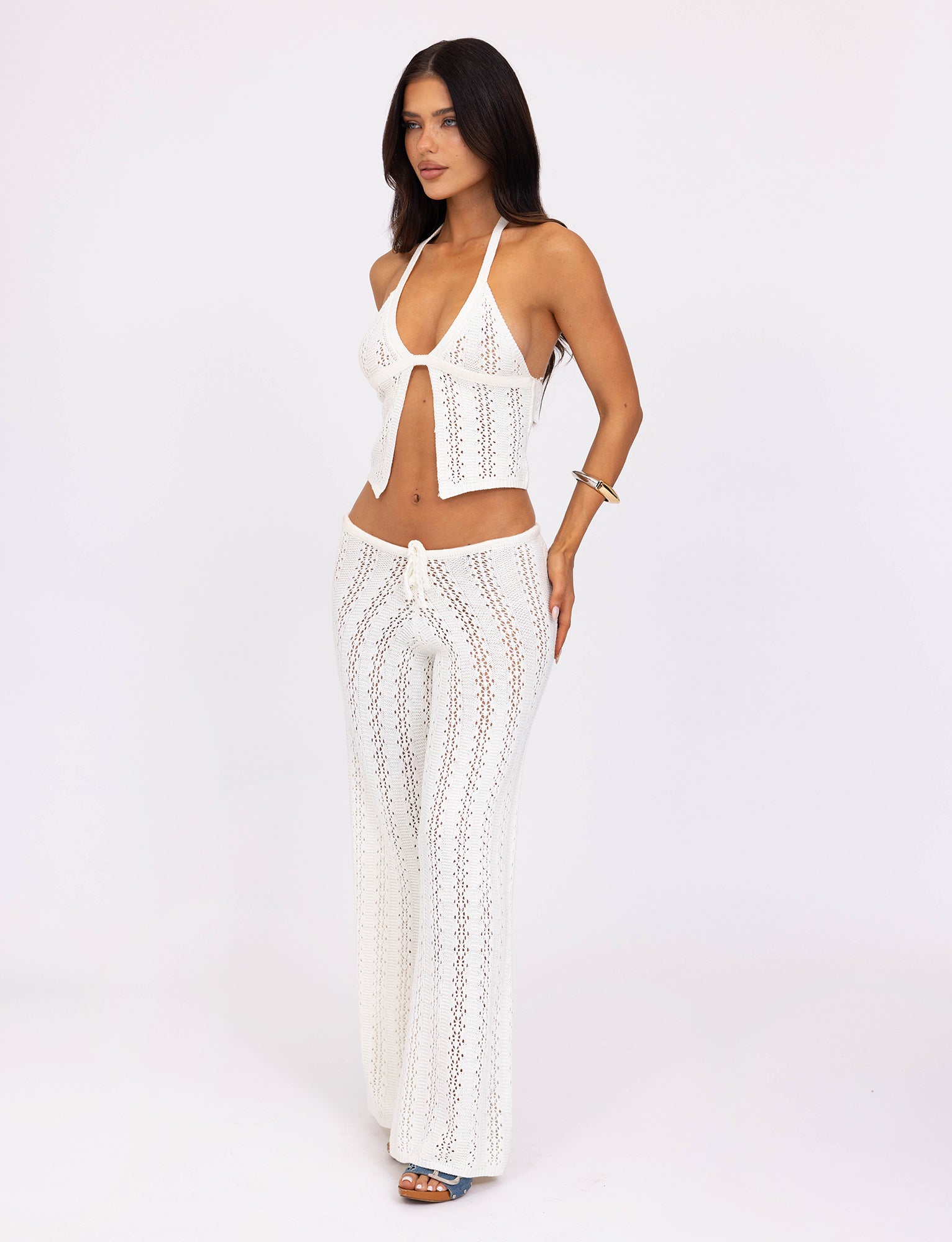 A woman with long, dark hair is wearing the white crochet halter top and matching SANDOR PANT - WHITE from TIGERMIST. She stands against a plain white background, posing with one hand on her hip and the other at her side. She accessorizes with a bracelet and blue sandals.