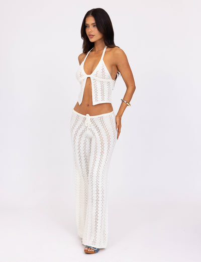 A woman with long, dark hair is wearing the white crochet halter top and matching SANDOR PANT - WHITE from TIGERMIST. She stands against a plain white background, posing with one hand on her hip and the other at her side. She accessorizes with a bracelet and blue sandals.