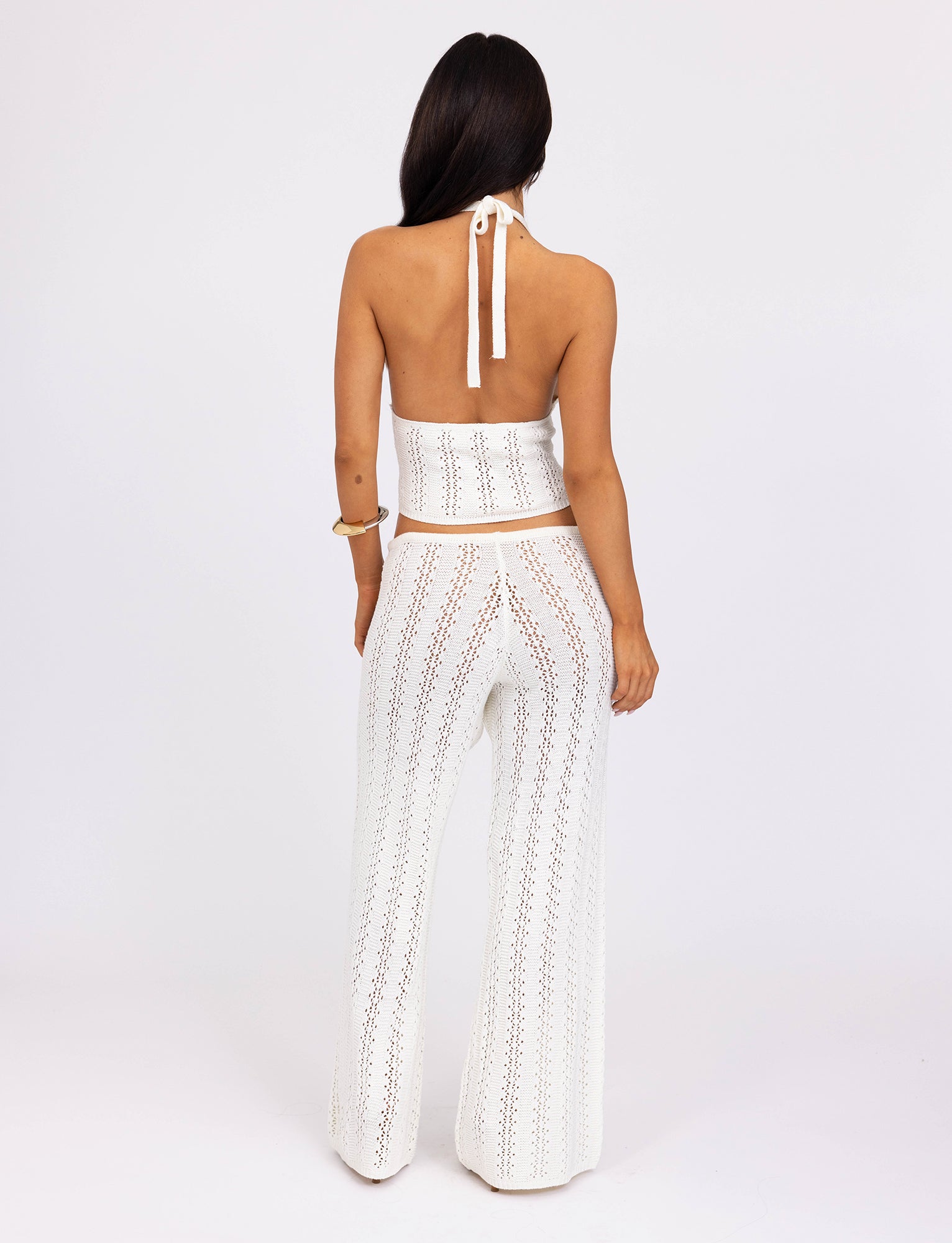 A person with long dark hair, seen from the back, wears a white crochet halter top and the SANDOR PANT - WHITE from TIGERMIST. The top ties around the neck, and the sheer pants reveal a light-colored undergarment. The background is white.