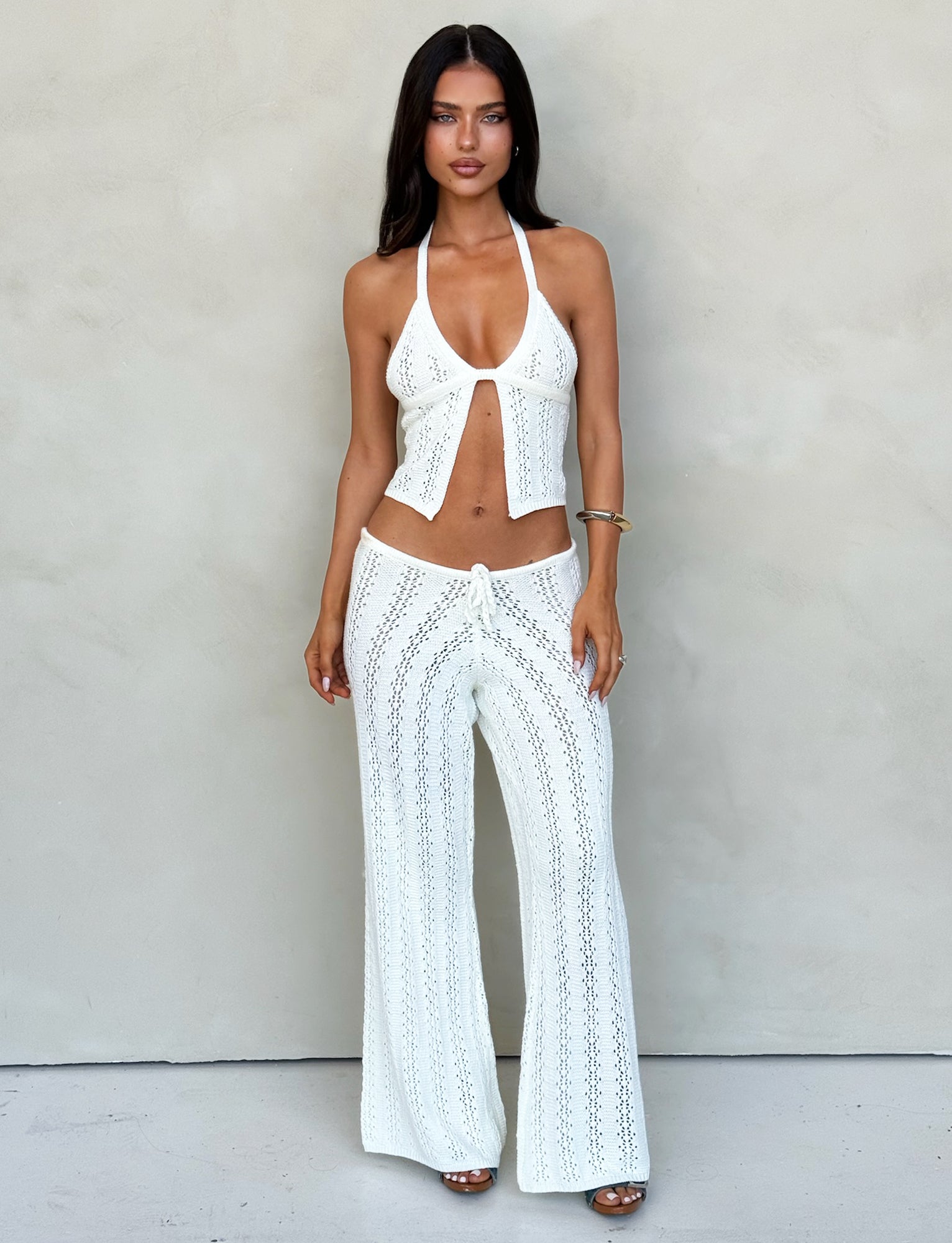 A woman stands in front of a neutral background wearing TIGERMIST's matching white crochet halter top and SANDOR PANT - WHITE. The halter top has an open front and ties at the neck, while the pants feature an intricate crochet pattern. She's accessorized with a bracelet.