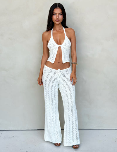 A woman stands in front of a neutral background wearing TIGERMIST's matching white crochet halter top and SANDOR PANT - WHITE. The halter top has an open front and ties at the neck, while the pants feature an intricate crochet pattern. She's accessorized with a bracelet.
