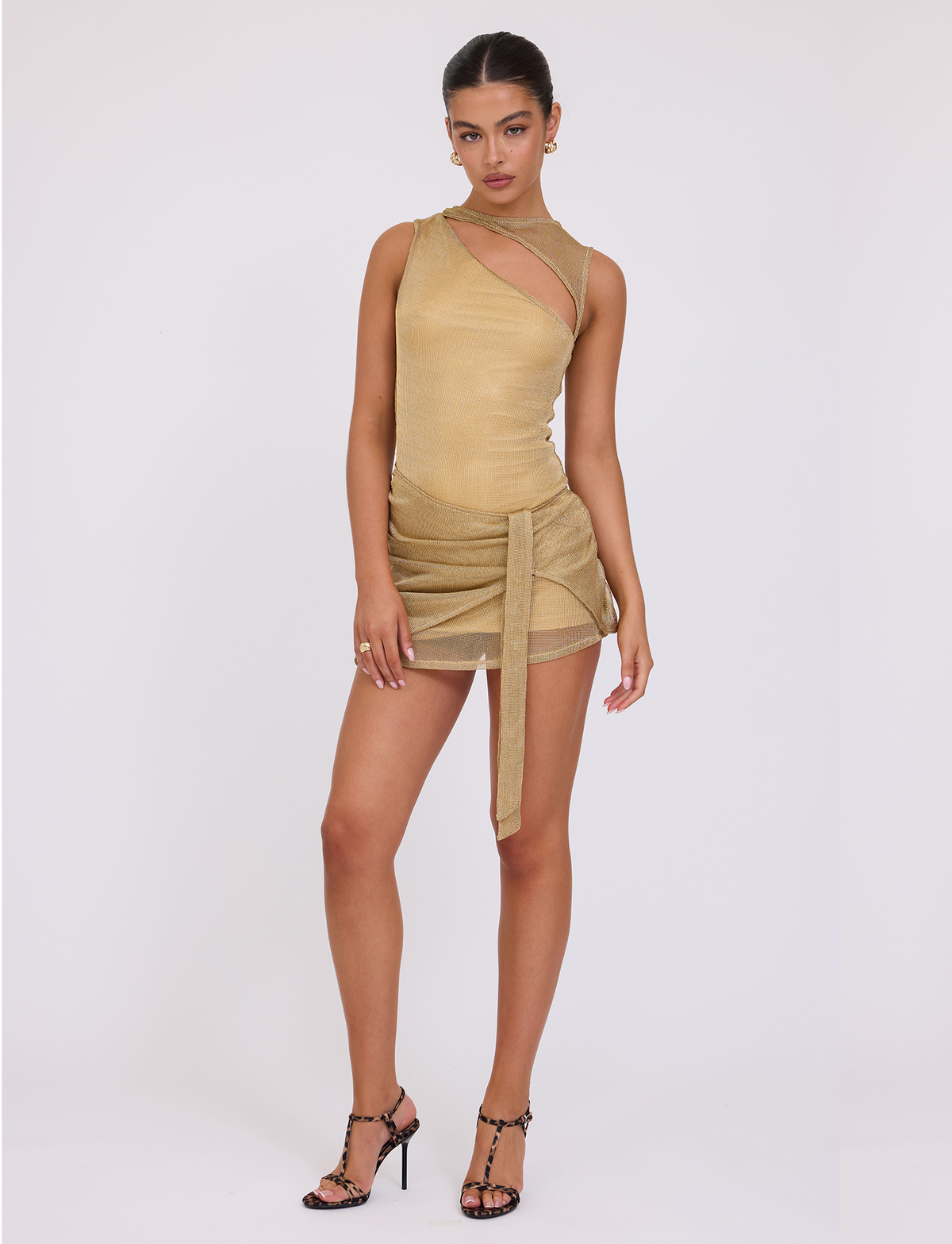 A woman stands confidently against a white background in the TIGERMIST AZELMA DRESS - GOLD, a sleeveless metallic knit mini with an asymmetric bust cutout and sash waist, styled with black strappy heels.