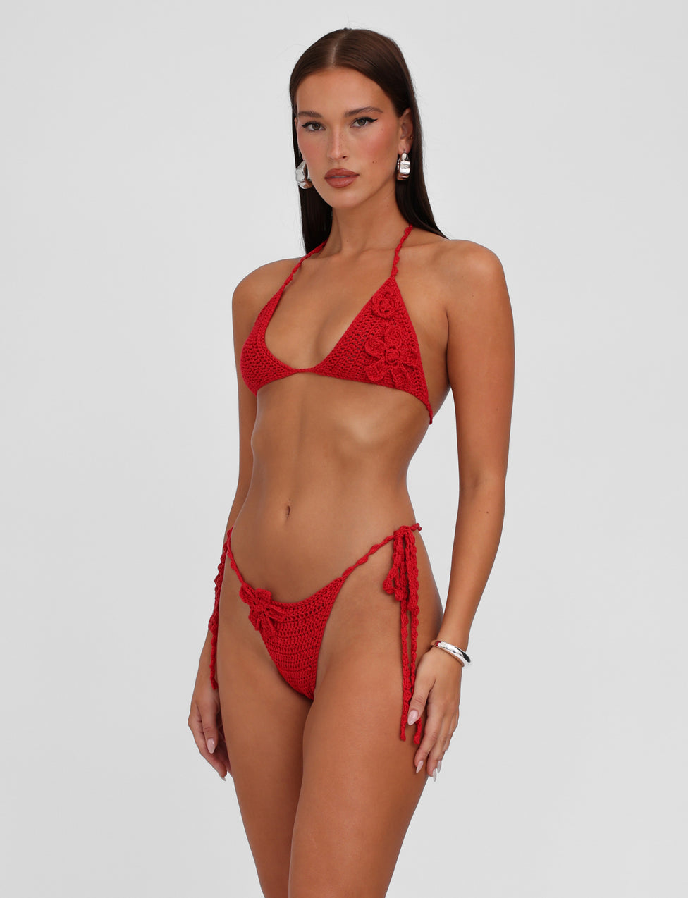 A woman is posing in a TIGERMIST MELIORA BIKINI BOTTOM in Berry Red with a neutral expression. The bikini features detailed patterns and wearable crochet, paired perfectly with silver hoop earrings and a bracelet. She has long brown hair and stands against a plain white background.