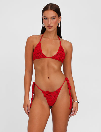 A woman in a Berry Red MELIORA BIKINI BOTTOM from TIGERMIST poses against a plain white background, showcasing the cheeky cut two-piece swimwear with tied sides on the bikini bottom and a halter top. She has long, straight hair and is accessorized with large hoop earrings, epitomizing wearable crochet elegance.
