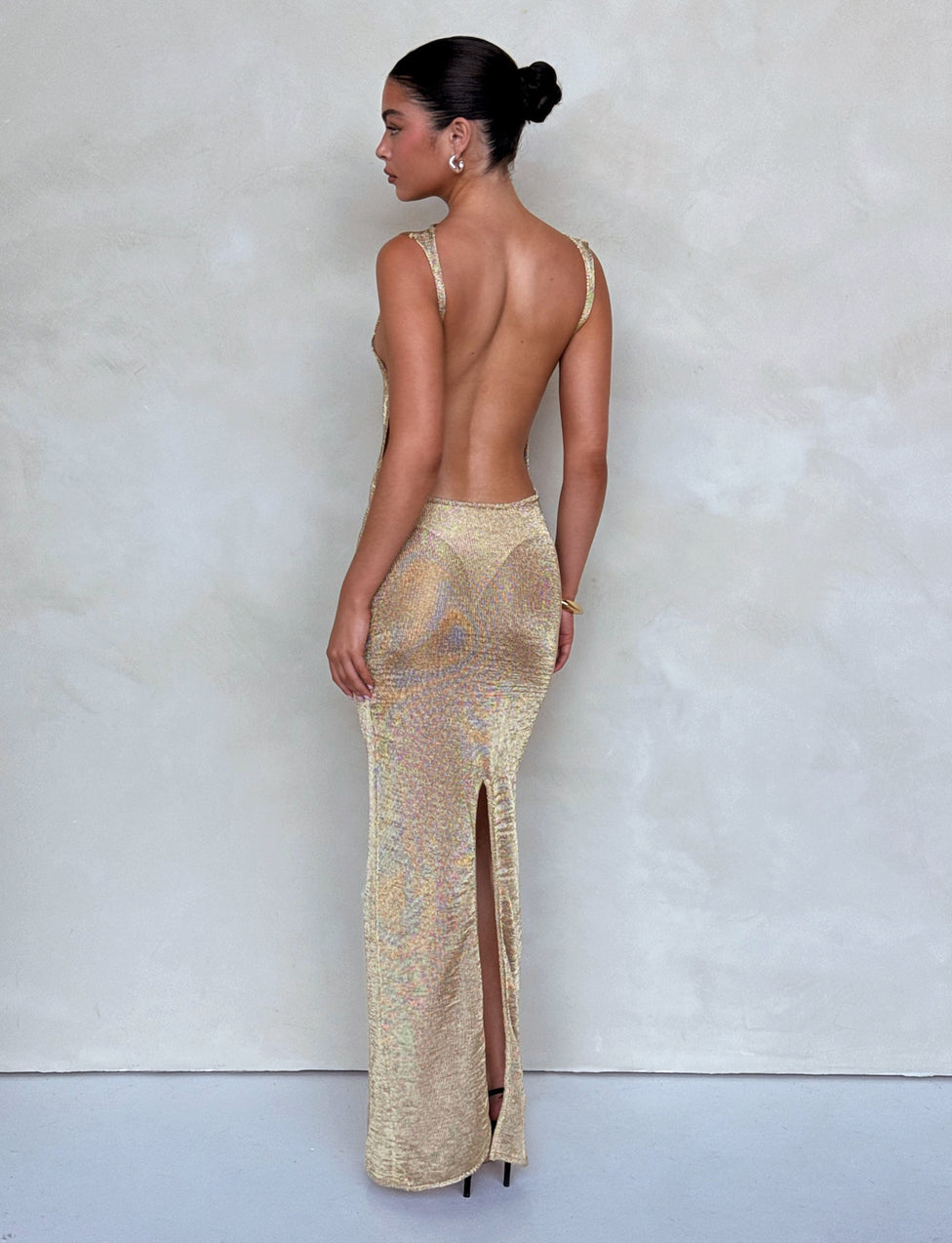 A woman stands in front of a plain background wearing the stunning ZAELA DRESS - GOLD from TIGERMIST. The backless, sleeveless dress features a shimmering gold fabric with a high slit at the back. With her hair styled in a neat bun and paired with high heels, she turns slightly to the side, showcasing the exquisite details of her dress.