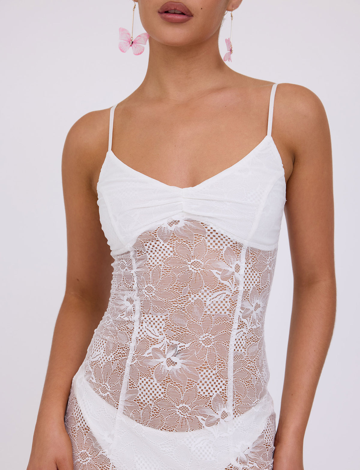 A woman is wearing the SASKIA DRESS in white from TIGERMIST, which features an intricate floral lace design and thin spaghetti straps. She is accessorizing with pink butterfly earrings. The image is cropped to show her from the chin to the upper thighs.