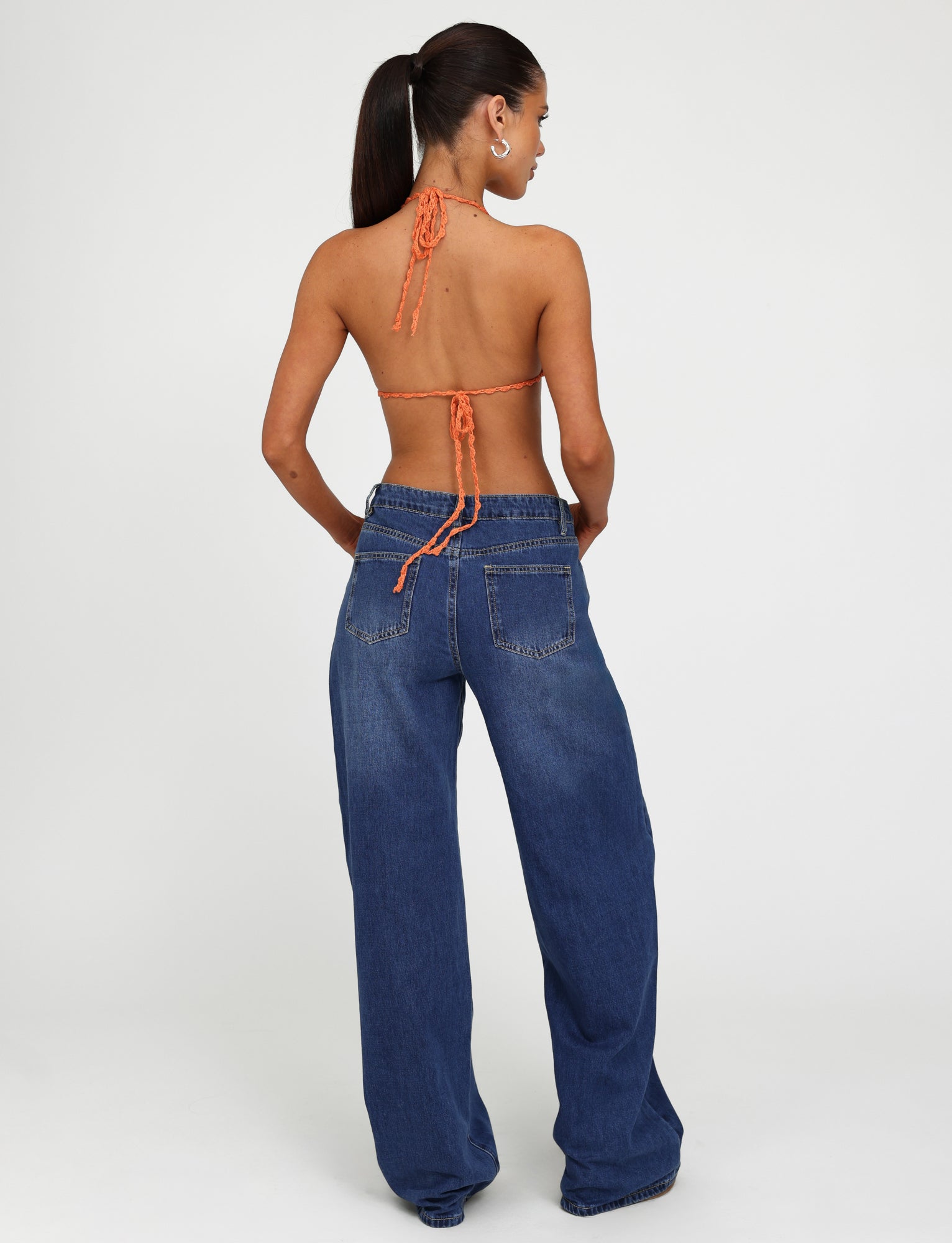 A woman with long dark hair tied in a ponytail is standing with her back to the camera. She is wearing a backless orange halter top from TIGERMIST and AVEREE JEAN - BLUE : DARK WASH wide-leg jeans. The background is plain white.
