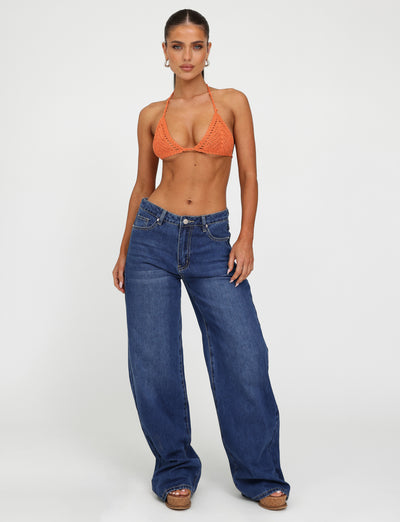 A person stands confidently against a white background, modeling a vibrant crocheted bikini top from Tiger Mist paired with the brand's dark wash AVEREE JEAN in blue. They accessorize with earrings and platform sandals, and their hair is styled back to highlight their features.