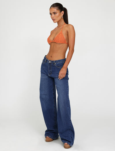 A woman stands against a plain white background, wearing a small orange crocheted bikini top from Tiger Mist and the Tigermist AVEREE JEAN - BLUE : DARK WASH. She has her hair tied back in a long ponytail and is looking off to the side. She is also wearing platform sandals.
