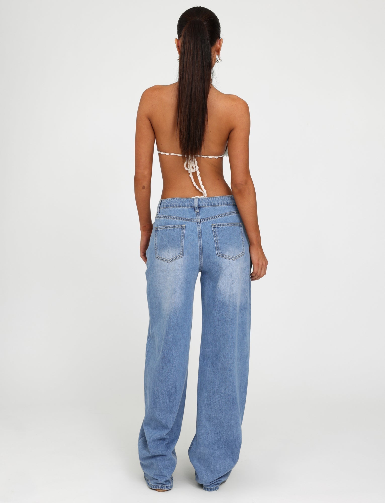 A woman is standing with her back to the camera, wearing a light-colored strappy top from Tiger Mist and the AVEREE JEAN - BLUE : MID WASH high-waisted, wide-leg jeans by TIGERMIST. Her long brown hair is tied in a low ponytail. The background is plain white.