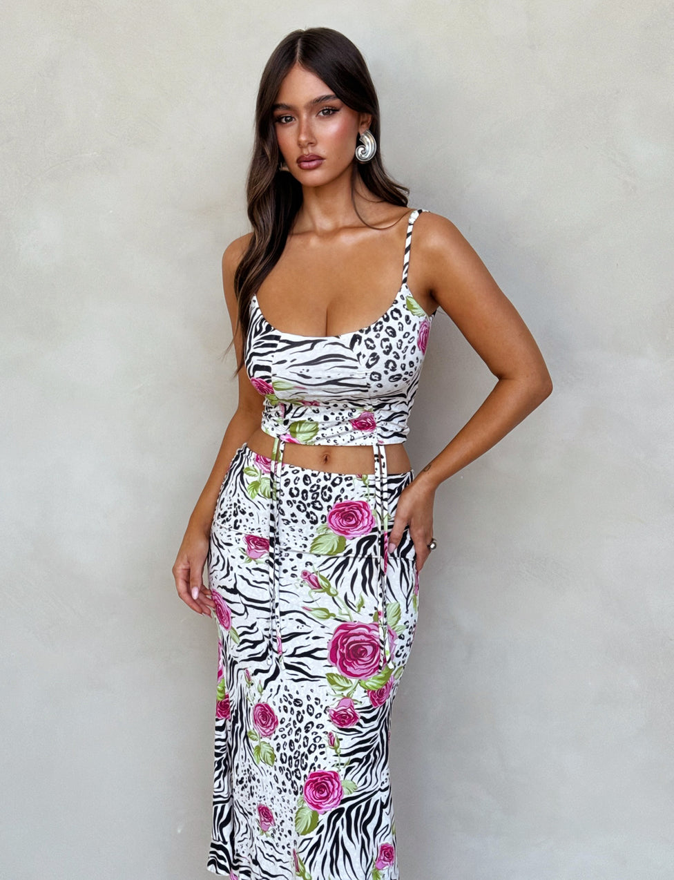 A woman with long, dark hair is wearing the IRIE TOP - MULTI : ZEBRA from TIGERMIST. The top features a fitted, animal print design with pink roses, thin spaghetti straps, and midriff cutouts. She is standing in front of a beige textured wall and looking directly at the camera.