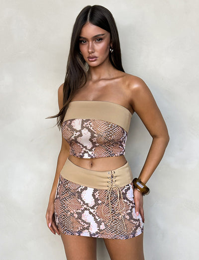 A woman stands against a light beige wall, wearing the FALLON SKIRT - BROWN : SNAKE co-ord set from TIGERMIST. The outfit consists of a strapless crop top and a matching mini skirt with lace-up detailing. She has long, dark hair and is accessorized with a bracelet.