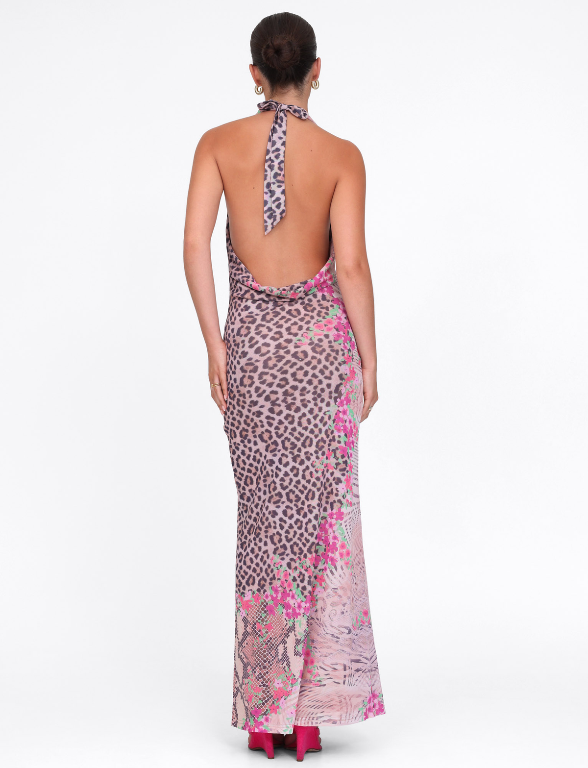 A woman with her hair in a bun wears the TIGERMIST CATALINA MAXI DRESS - NEUTRAL: ANIMAL FLORAL BORDER, a backless halter-style, floor-length dress, paired with pink heels against a plain white background.