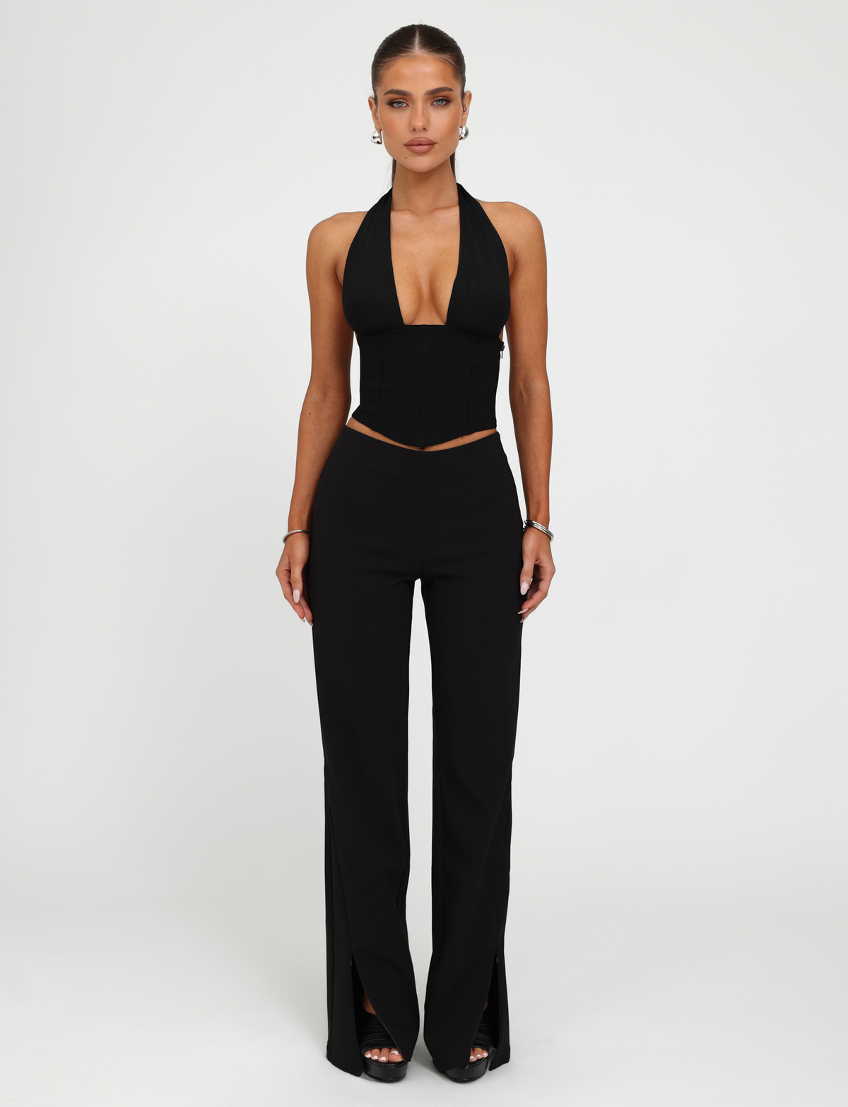 A woman stands against a white background wearing the TIGERMIST JANECE TOP in black, a corset-style halter-neck with a deep V neckline, paired with high-waisted black pants with front slits. Her hair is tied back and she accessorizes with earrings and bracelets.