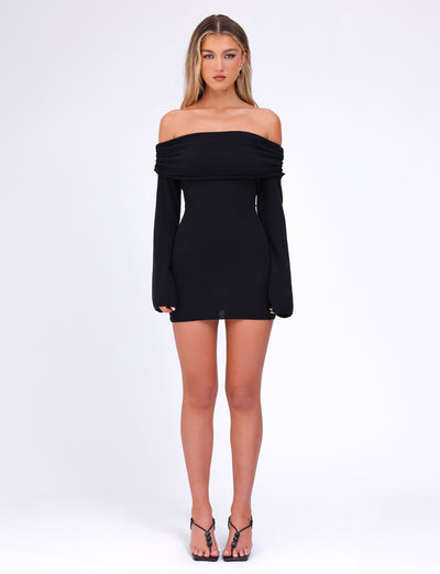 A woman stands against a plain white background, wearing the off-the-shoulder, long-sleeved DIEDRE JUMPER DRESS - BLACK by TIGERMIST, which features a fitted silhouette. She completes her outfit with black strappy heeled sandals. She has long, straight hair and a neutral expression.