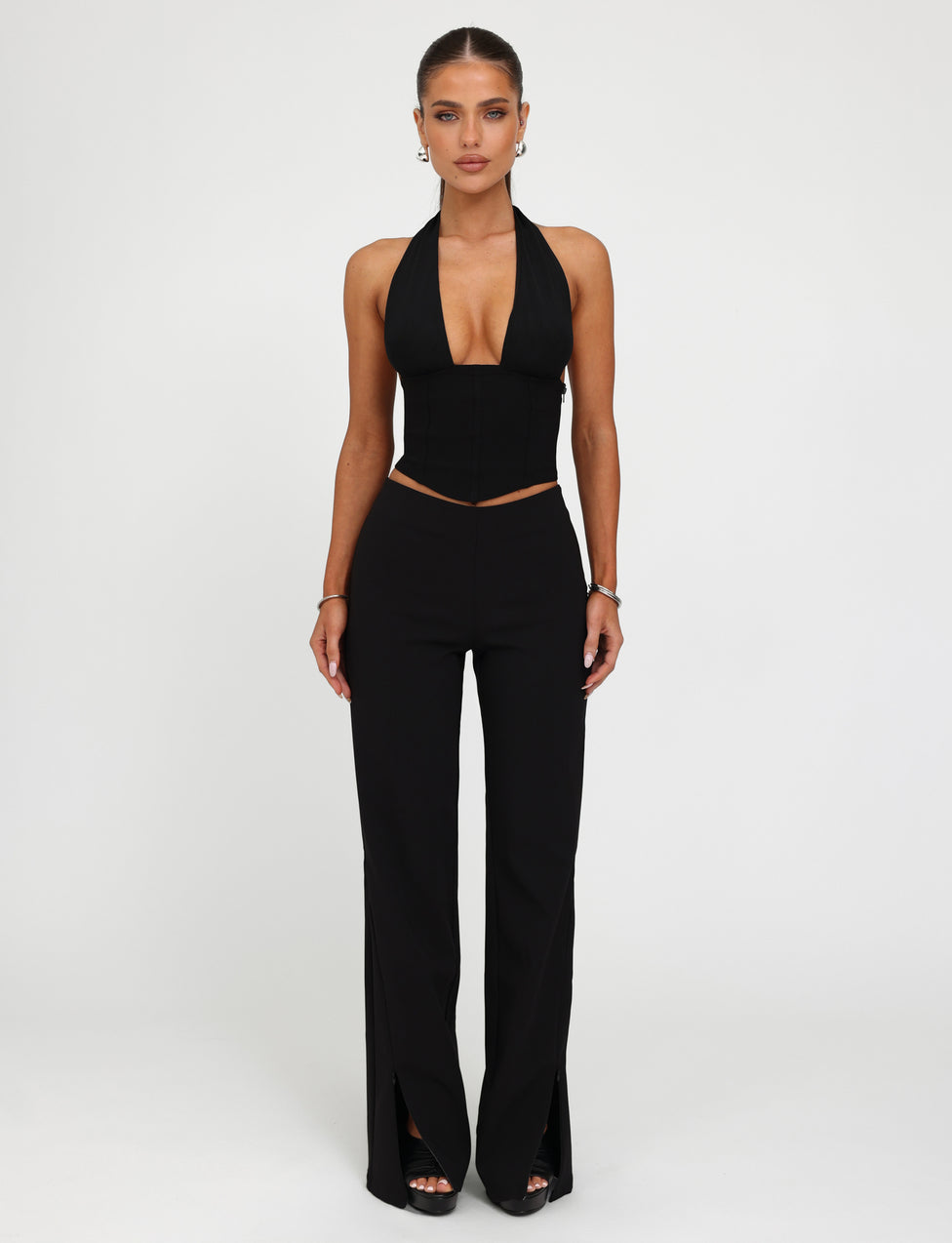 A woman stands against a plain white background. Sporting a sleek outfit from TIGERMIST, she wears a black halter top with a deep V-neckline and the DAMIRA PANT in black, which features high-waisted design and slits at the front hems. Her hair is pulled back, accessorized with small hoop earrings and a bracelet.