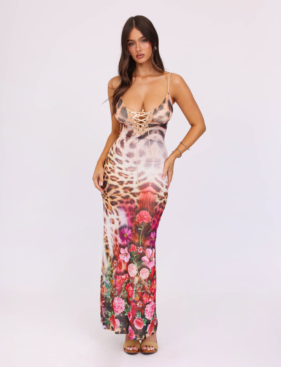 A woman with long dark hair stands against a plain background, wearing a colorful KALILAH DRESS - MULTI : LEOPARD by TIGERMIST. The maxi dress features a mix of animal and floral prints with lace-up details at the bust. She is posing with her hands lightly grasping the sides of her dress.