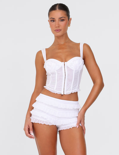 A woman stands confidently wearing the TIGERMIST MIKEL SHORT in white paired with a matching corset top. The outfit, showcasing a bodycon fit and ruffle details, exudes elegance through its delicate design. She poses effortlessly against a plain white background.