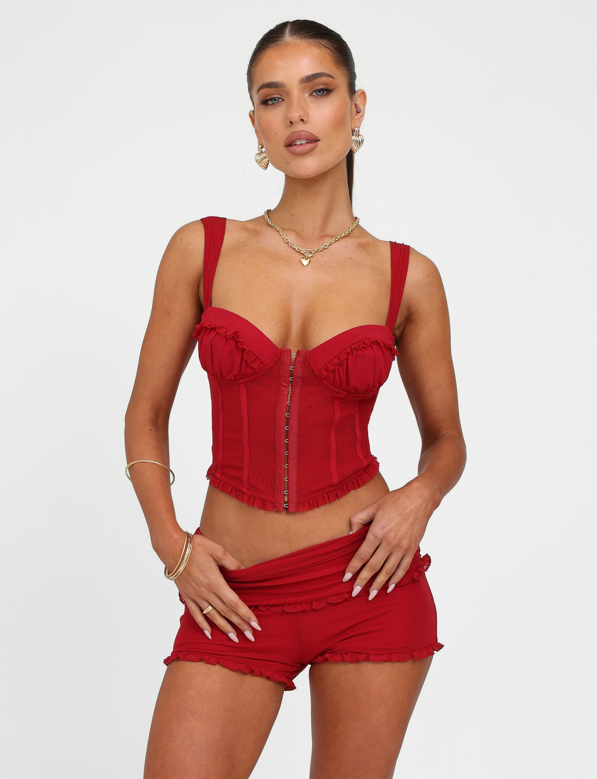 A woman poses against a white background wearing the TIGERMIST GINNY TOP - RED : CHERRY RED, a corset-style piece with a sweetheart neckline and front zipper, paired with matching red shorts, gold jewelry, and her hair pulled back.