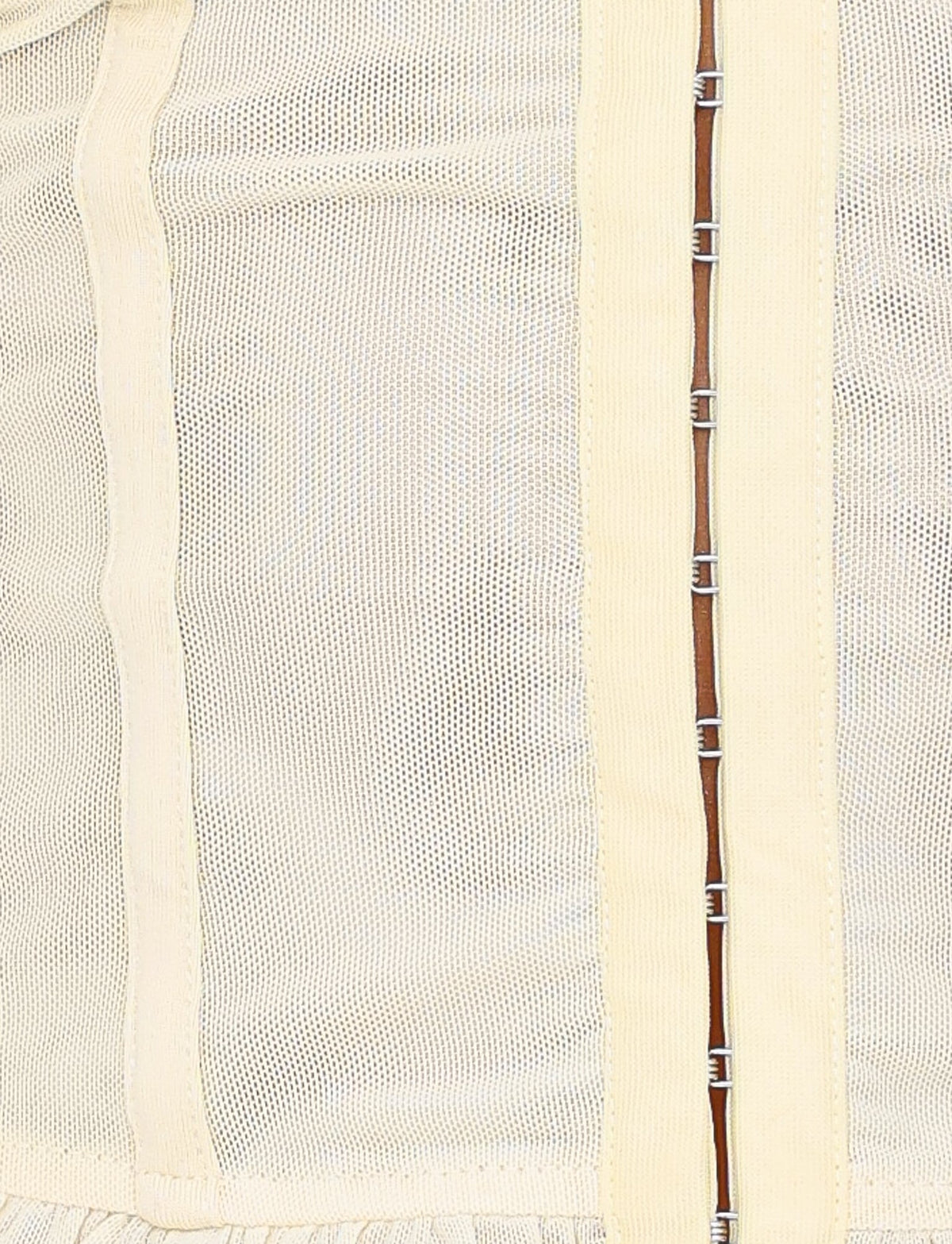 A close-up of a textured fabric in cream color, evocative of the Ginny Top from TIGERMIST. This fabric showcases vertical mesh panels and a line of small metal hooks running vertically, indicating a fastening mechanism typical for garments with a sweetheart neckline or bodycon design.