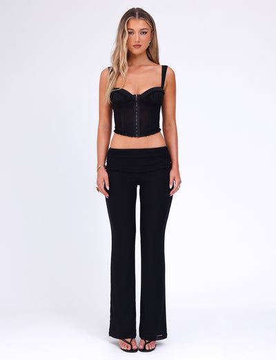 A woman with long blonde hair stands against a plain white background. She is wearing the GINNY TOP - BLACK from TIGERMIST, which is a black, sleeveless crop top featuring a zipper in the front and a sweetheart neckline. She pairs it with black flared pants and black strappy heels. The bodycon silhouette of her outfit emphasizes her figure as she keeps a neutral expression with her arms relaxed at her sides.