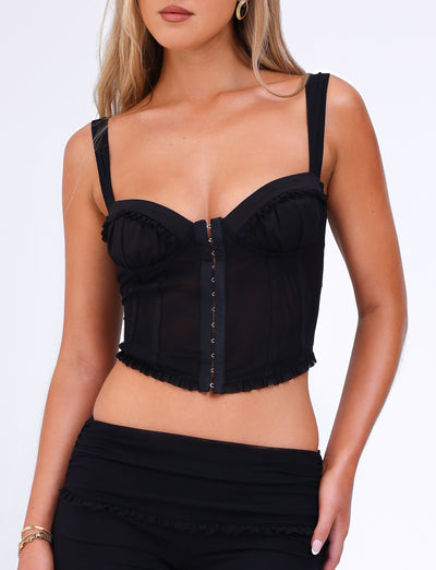 A woman is wearing the GINNY TOP - BLACK by TIGERMIST, featuring thin straps, ruffled detailing, and a sweetheart neckline that highlights its bodycon silhouette. She pairs it with matching high-waisted black pants. The minimalistic background accentuates the outfit beautifully.