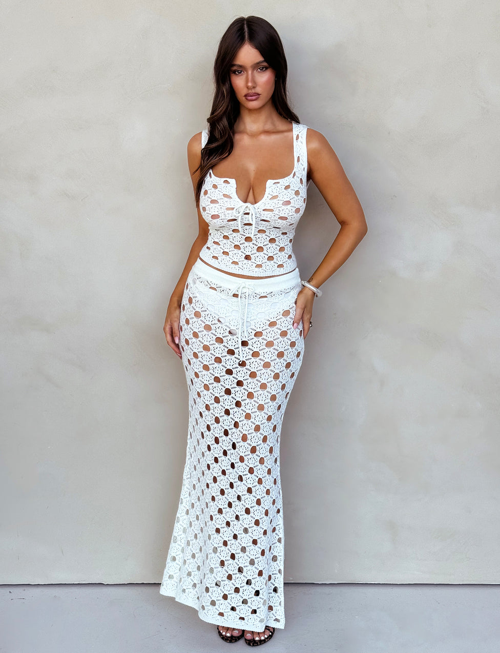 A woman stands against a neutral background wearing the TIGERMIST KINCADE TOP in white, part of a matching crochet set that includes a cropped top and long skirt. She has long, dark hair and poses with her hands on her hips, looking directly at the camera.