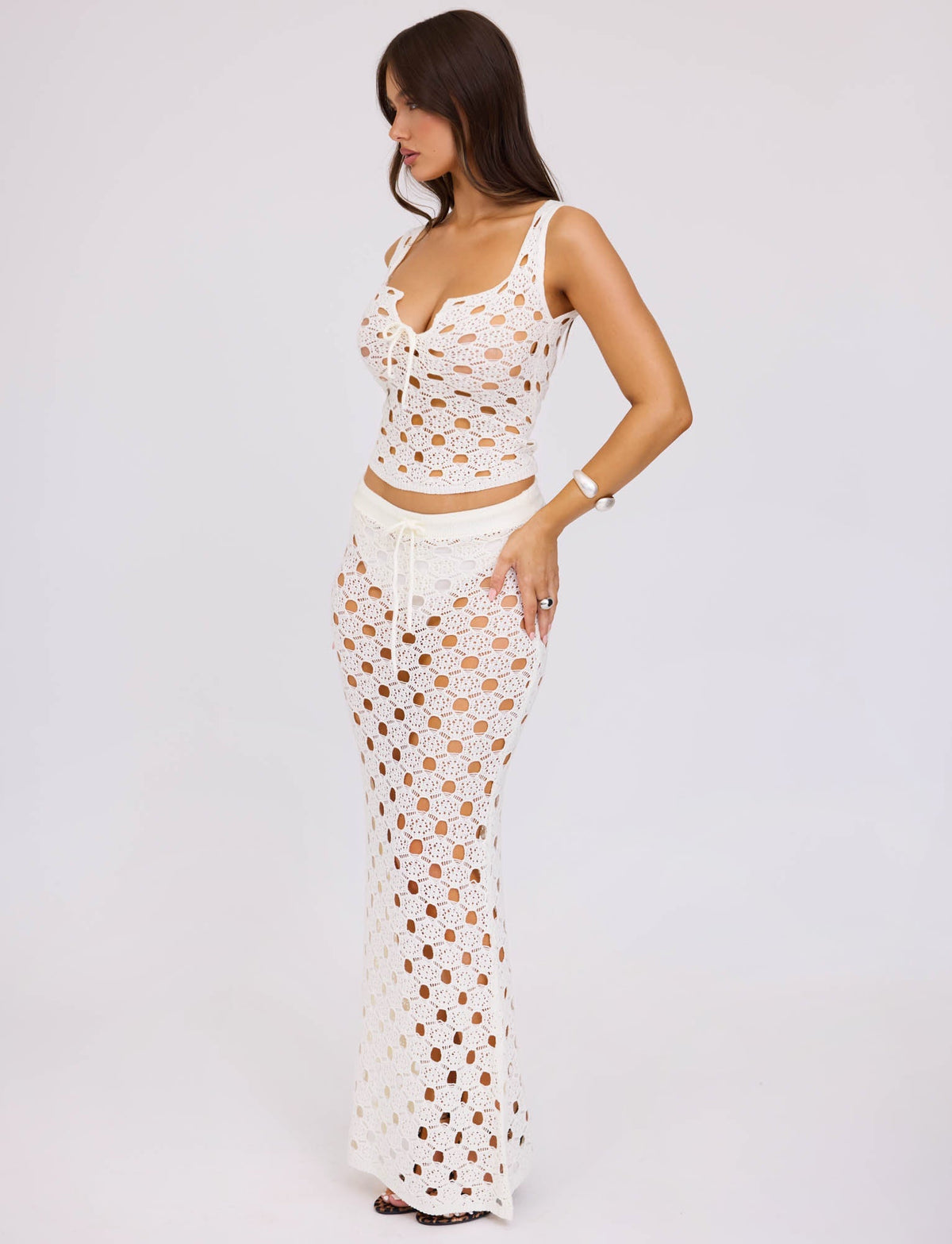 A woman with long brown hair is wearing the KINCADE TOP - WHITE from TIGERMIST. The top has an intricate crocheted design exposing her skin underneath. She stands against a plain white background, looking to her right with one hand on her hip.