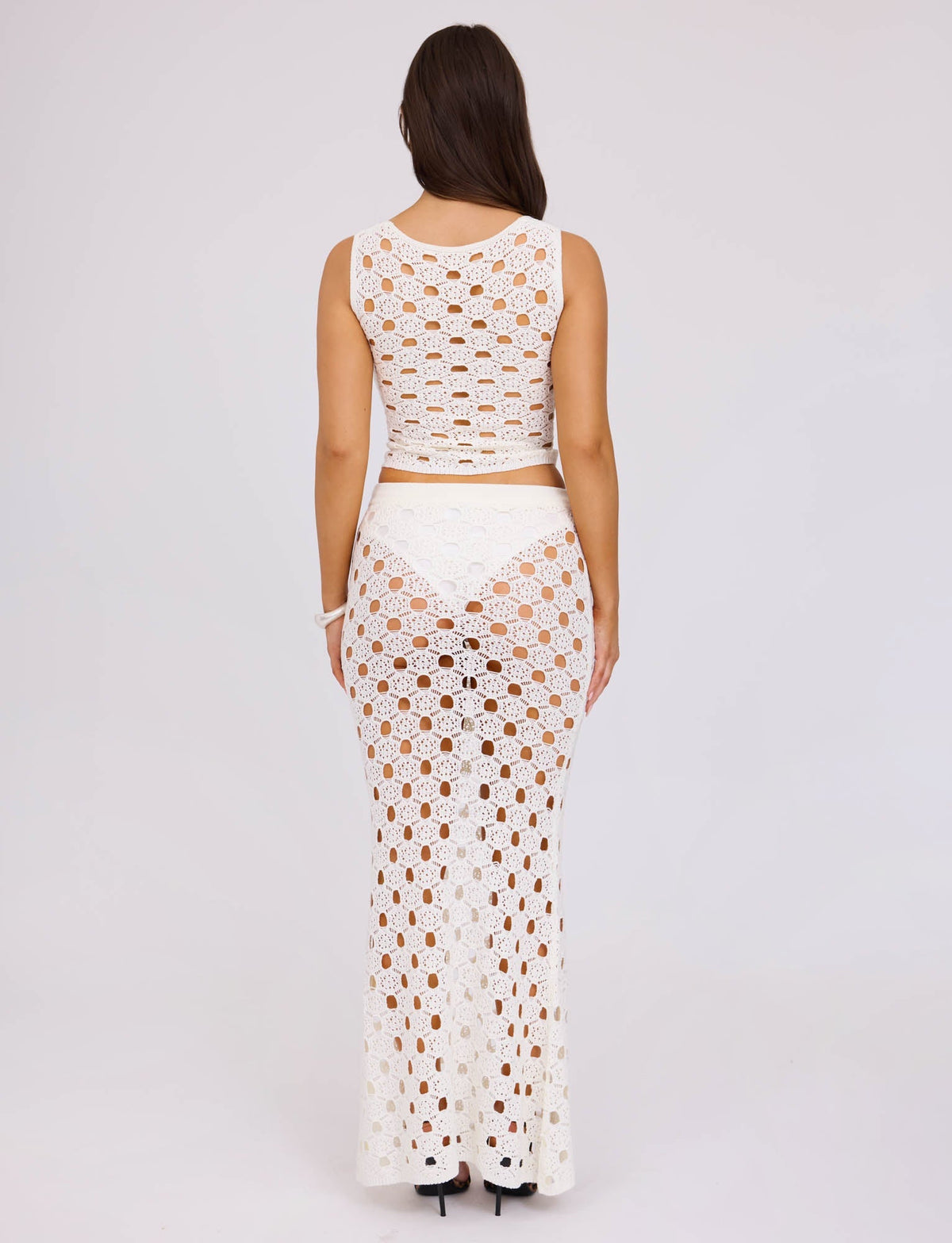 A woman stands facing away, wearing the Kincade Top in white from TIGERMIST, paired with a matching long skirt. The outfit features an intricate see-through crochet pattern. She has long brown hair and is wearing black high-heeled shoes against a plain, light-colored background.