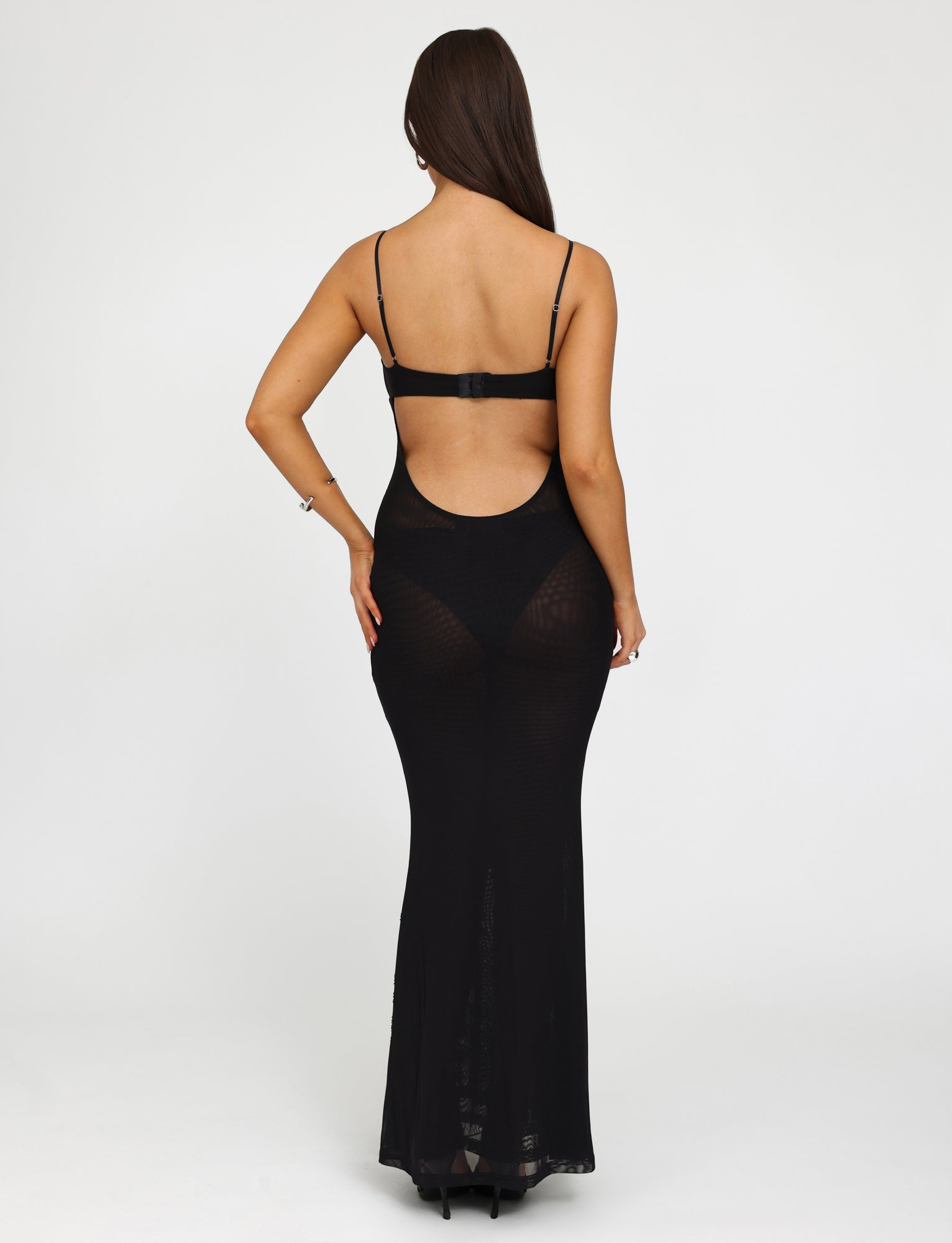 A person stands facing away from the camera, showcasing the TIGERMIST OCTAVIA DRESS - BLACK, a long, form-fitting dress with thin straps and an open back. The background is plain white, highlighting their long, straight brown hair cascading down their back.